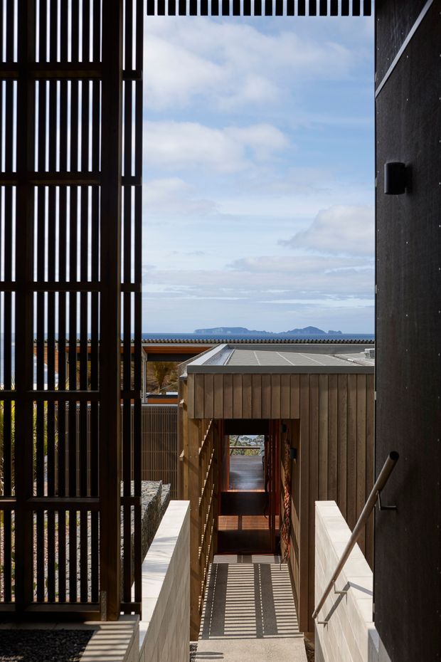 A narrow architectural passage and stairway framed by dark vertical timber screens and a dark rendered wall, leading down toward a timber-clad lower structure. Strong linear shadows fall across the stairs and walkway. Beyond the roofline, blue sky and distant mountainous or coastal landforms are visible. The composition emphasises layered thresholds, privacy screening, natural timber, and a dramatic framed outlook.