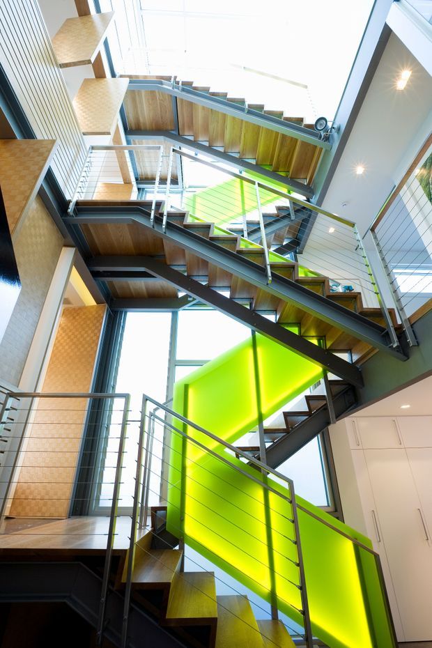 A dramatic multi-level interior stairway viewed upward through a tall, bright stairwell. Multiple zigzagging timber-tread stair flights are supported by dark steel stringers and enclosed by slender horizontal metal cable railings. Large floor-to-ceiling glazing brings in abundant daylight, while vivid translucent lime-green panels form striking balustrade or stairwell feature surfaces. Timber wall and ceiling elements, white cabinetry, and recessed ceiling lights reinforce the contemporary architectural character.