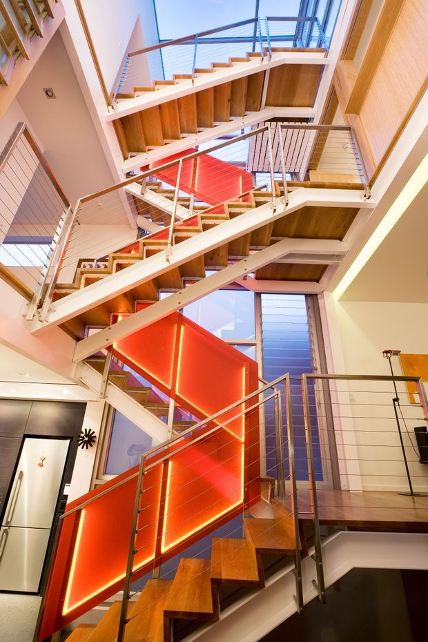 A dramatic multi-level interior stairway viewed upward through an open vertical stairwell. Several flights of timber-tread stairs overlap around a central void, with slim metal and horizontal cable balustrades. Warm timber soffits and treads contrast with white structural stringers, pale walls, extensive glazing, and a vivid red illuminated feature wall beneath the stairs.