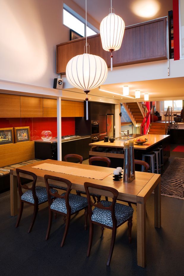 A warm, eclectic open-plan dining and kitchen interior with a timber dining table and upholstered dining chairs in the foreground. The space features extensive dark and mid-tone timber built-in cabinetry, a vivid red backsplash, black kitchen joinery, a central island, sculptural pendant lights, and a visible staircase leading to an upper mezzanine or landing. High-level glazing and layered architectural volumes create a loft-like atmosphere.