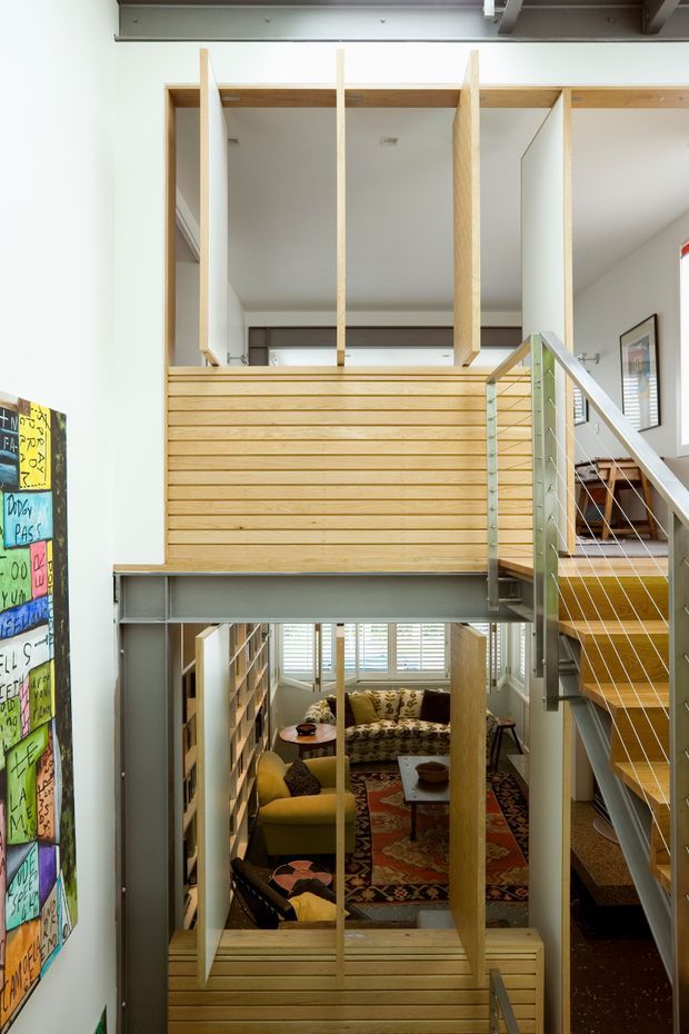 Vertical interior view of a compact multi-level home with an open stairway and mezzanine. Exposed grey steel beams frame timber slat balustrades and partition screens, while a lower living room with sofas, patterned rug, shelving, and large windows is visible below. The space has an industrial-modern character with warm timber detailing and strong vertical connections between levels.