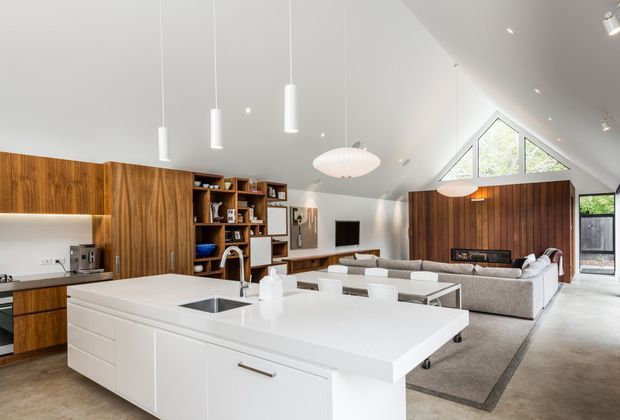 Bright open-plan kitchen and living room with a large white island, integrated sink, timber cabinetry and open shelving. The adjoining living area has a large sectional sofa, media wall and fireplace, while a dramatic pitched ceiling with triangular clerestory glazing and pendant lighting adds architectural interest.