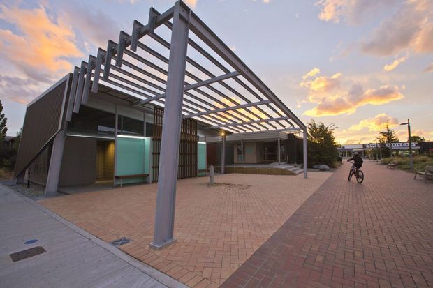 A contemporary low-rise commercial building viewed from an outdoor paved plaza at sunset. The building has dark vertical cladding, extensive glazing, and a prominent steel pergola or framed canopy projecting over the brick-paved forecourt. A bicycle and surrounding site elements are visible, with warm interior lighting beneath the canopy.