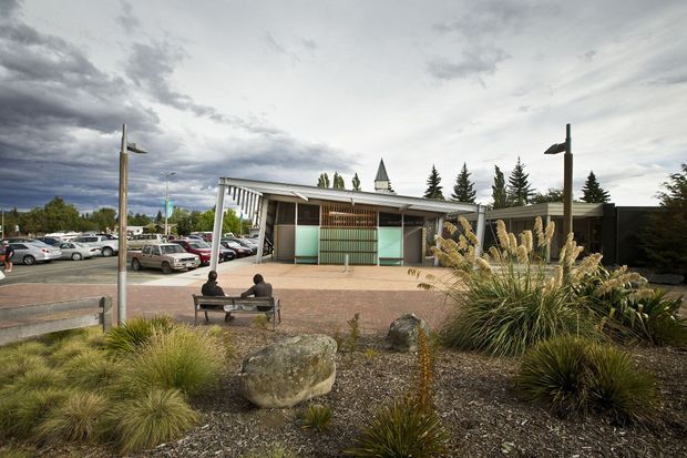 Exterior view of a low-rise contemporary public or commercial building, set within a landscaped forecourt. The building has a long, low roofline, extensive glazing, vertical timber-like screening, pale rendered or concrete walls, and a small projecting entrance canopy. Native grasses, boulders, benches, and modern pole lighting occupy the foreground, with a car park visible behind. The overcast sky gives the scene a subdued, slightly dramatic atmosphere.