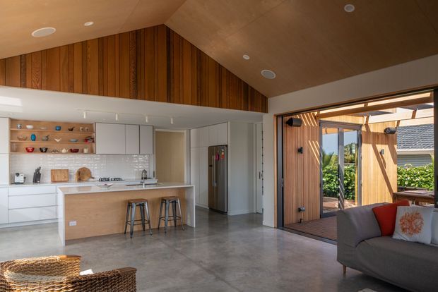 Open-plan contemporary kitchen and living area with a large island, white integrated cabinetry, timber-lined vaulted ceiling and upper wall, polished concrete flooring, and wide glazing opening to a timber-clad outdoor courtyard. A grey sofa and woven seating occupy the living zone.