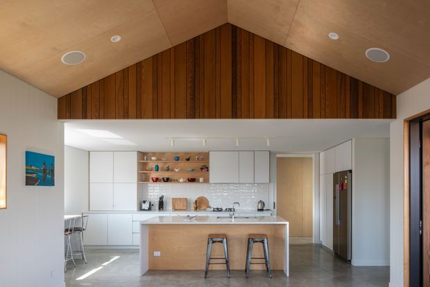 Contemporary open-plan kitchen viewed from the living area, featuring a large central island with timber panelled base, white built-in cabinetry, pale tiled splashback, stainless-steel appliances and stools. A dramatic pitched ceiling is lined with light timber, while the gable end has dark vertical timber panelling. Polished concrete flooring and recessed ceiling lights complete the clean, warm interior.
