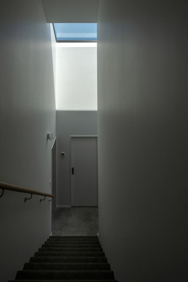 A narrow, tall stairwell viewed from the upper stairs, with dark carpeted steps descending toward a grey landing and closed door. Smooth pale grey walls rise to a high ceiling with a rectangular skylight that brings in cool natural light. A timber handrail with dark metal brackets runs along the left wall, creating a minimalist, subdued interior.