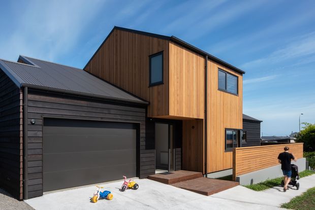Exterior view of a contemporary multi-storey house with a bold timber-clad upper volume, dark horizontal cladding on the garage and lower sections, a large charcoal sectional garage door, and a small timber entry deck and privacy fence. The home sits along a residential driveway and walkway under a clear blue sky.