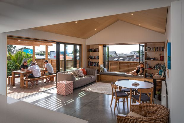 Bright, open-plan contemporary living and dining area with a pitched timber-lined ceiling, polished concrete floor, extensive timber-framed glazing, built-in bookshelves, and a window seat. The room opens to a timber deck where people are dining, with residential rooftops and greenery visible outside.