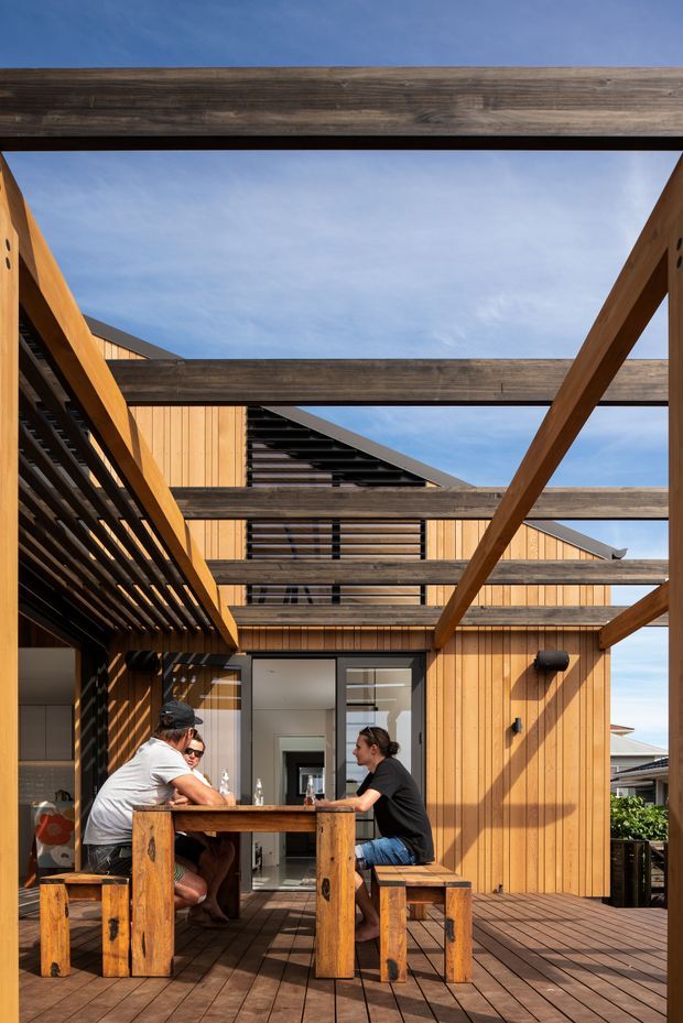 A sunlit timber-clad outdoor courtyard or deck framed by substantial exposed timber beams and pergola elements. A built-in timber dining table with benches occupies the foreground, while a multi-level timber-clad building with open-air voids, horizontal slatted screening, dark roof planes and glazed doors forms the background. The image emphasizes exposed structure, warm natural wood, and indoor-outdoor living.