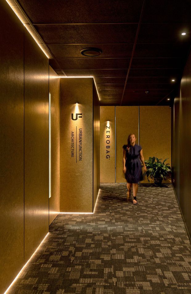 A dimly lit commercial office entry corridor with bronze-toned wall and ceiling panels, integrated linear and recessed lighting, patterned carpet, and illuminated architectural signage for Urban Function Architecture and Zerobag. A potted plant and a person appear near the far end of the hallway. The space has a refined, atmospheric corporate lobby aesthetic.
