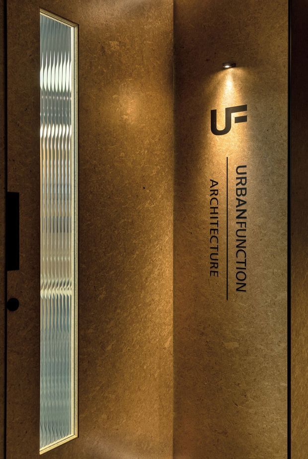 A dimly lit architectural interior featuring a textured bronze-brown wall with illuminated signage reading “URBAN FUNCTION ARCHITECTURE.” A tall narrow reeded or patterned glass window with a dark frame runs vertically along the left side, while a small wall-mounted spotlight highlights the logo and lettering. The image has a refined, dramatic commercial-studio or office reception character.