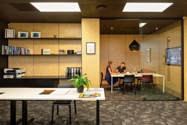 Contemporary commercial office interior with a collaborative meeting area, work tables, open shelving, storage cabinetry, carpeted flooring, wood-toned wall panels, and a suspended pendant light. A glazed meeting room and people working at a table are visible in the background.