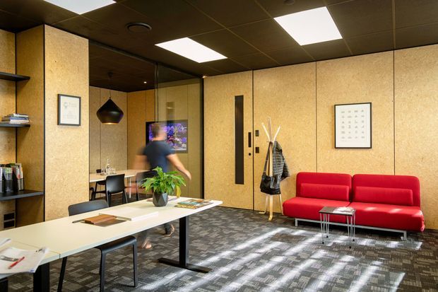 A compact contemporary office interior with pale wood-panelled walls, dark shelving and ceiling, integrated workstations, a red sofa, pendant lighting, framed artwork, coat storage and patterned carpet. Daylight enters through glazing, creating a professional yet inviting workspace.