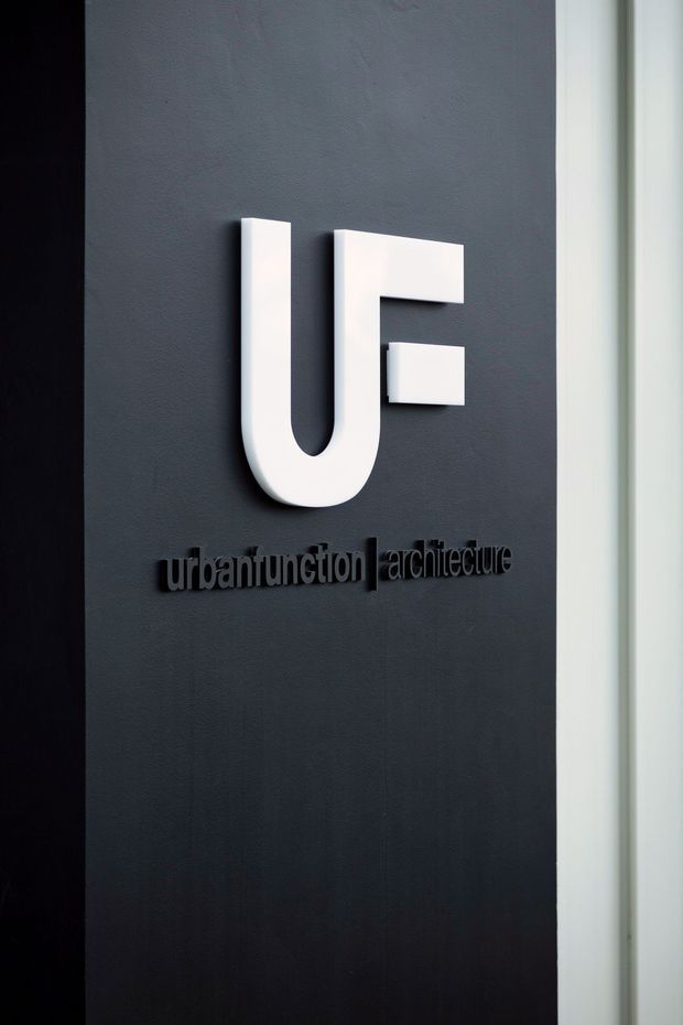 A close-up of a minimalist architectural studio or office sign mounted on a dark charcoal wall. A large white abstract “UF” logo sits above black raised lettering reading “urbanfunction | architecture,” with a narrow white doorframe or wall edge visible at the right.