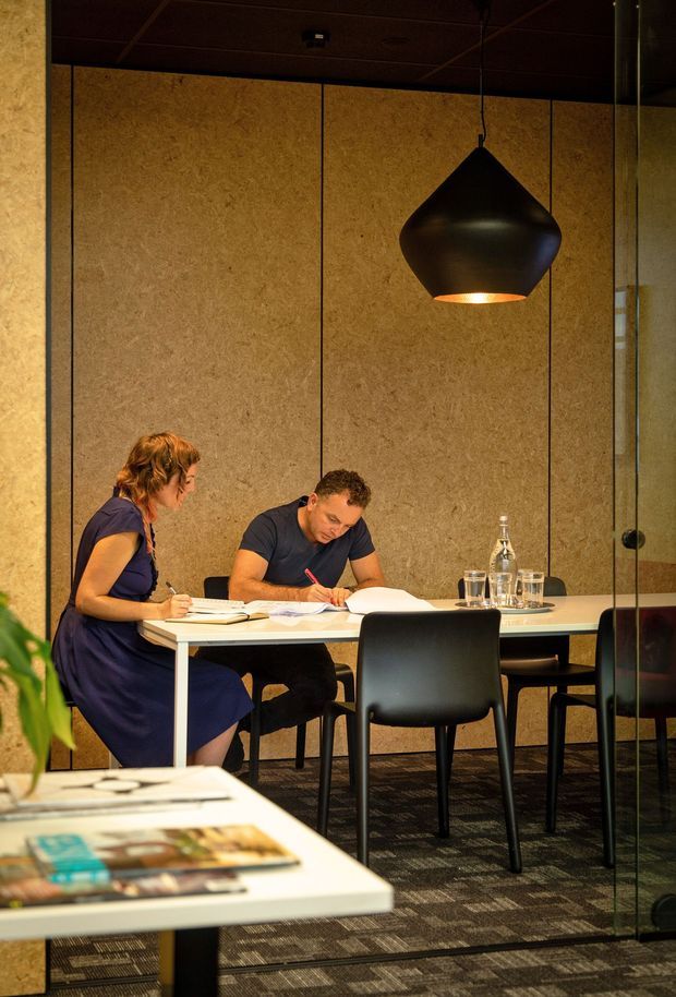 A warm, moody commercial interior showing two people seated at a white table in a meeting or collaborative workspace. The room features textured brown wall panels, dark ceiling panels, a large sculptural black pendant light, black chairs, glass partitions, and dark patterned carpet. A foreground table displays magazines or printed materials.