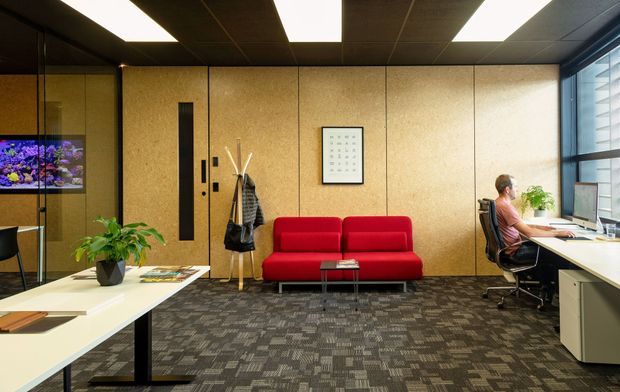 A contemporary office reception and waiting area with a vivid red sofa, patterned carpet, pale wood-toned wall panelling, a glass meeting room or aquarium feature at left, and workstations along the glazed right side. Linear recessed ceiling lights and black-framed glazing create a clean commercial interior.