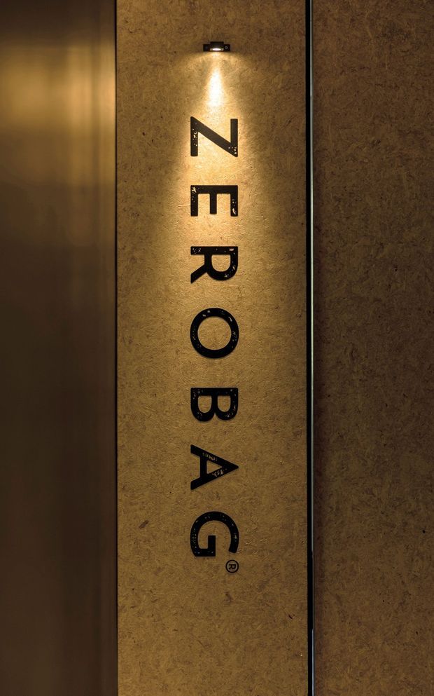 A close-up, vertically oriented commercial interior sign reading “ZERO BAG,” rendered in black raised lettering on a warm textured tan-brown wall panel. A small wall-mounted spotlight illuminates the sign from above, creating a moody, refined atmosphere. Dark vertical wall or panel edges frame the central sign.