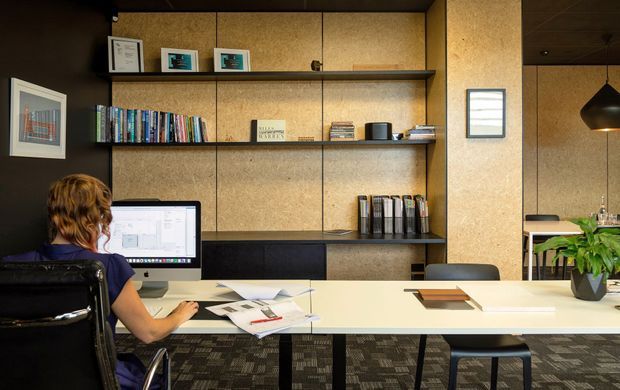 A contemporary home office or studio workspace with a long white shared desk, computer workstation, office chair, shelving filled with books and binders, framed artwork, and a large cork-textured built-in wall. A partial dining or breakout area with pendant lighting and plants is visible at right. The room has dark ceilings and patterned carpet tiles.