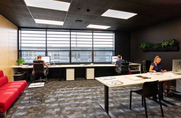 A contemporary open-plan commercial office with multiple workstations arranged along a large glazed window wall, a shared drafting or meeting table in the foreground, dark walls and suspended acoustic ceiling tiles. The space includes patterned carpet flooring, black-framed horizontal window blinds, modern office furniture, a red lounge sofa, and a wall-mounted planter.