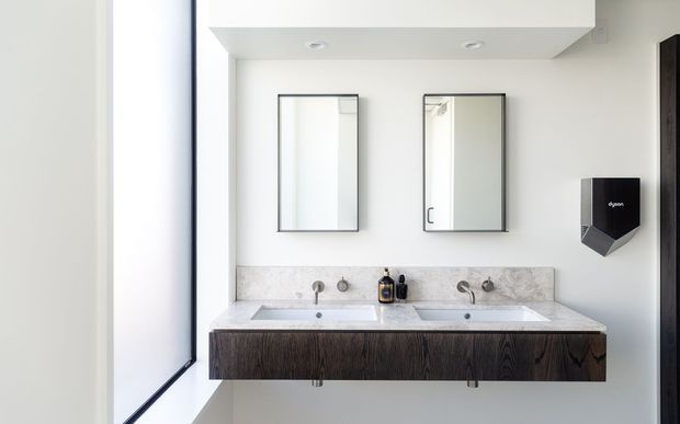 Bright contemporary bathroom with a double wall-mounted vanity, two integrated rectangular basins, dark wood-grain cabinetry, pale stone benchtop, twin mirrors, wall-mounted chrome tapware, and a black wall-mounted hand dryer. A large frosted or translucent window provides soft natural light.