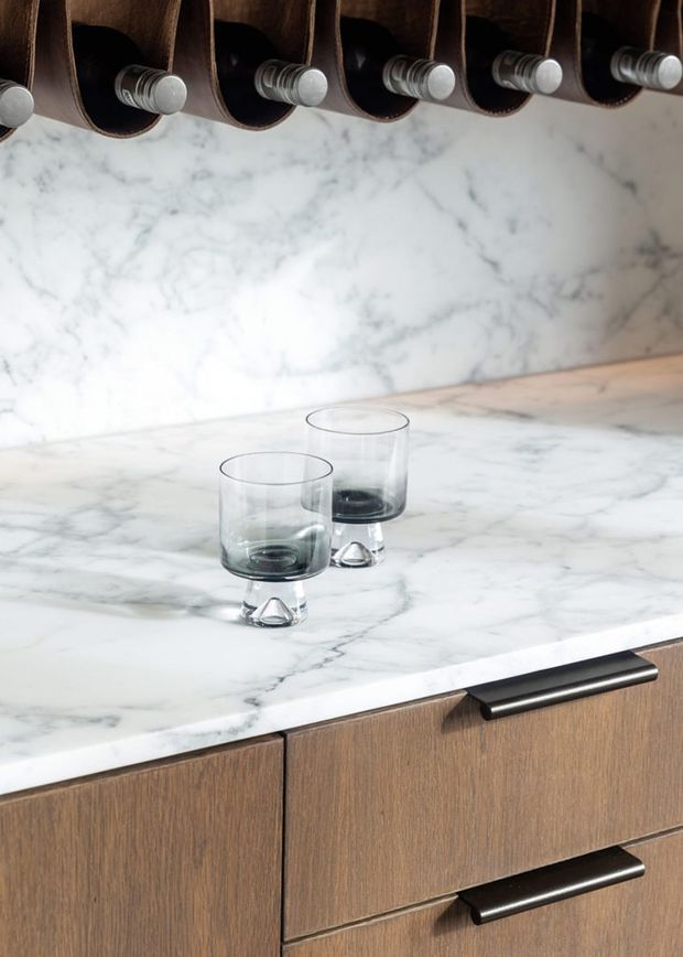 Close-up of a refined kitchen or bar joinery detail featuring a white veined marble benchtop and matching splashback, warm mid-tone timber cabinetry, and dark metal handles. Two clear drinking glasses sit on the stone surface, while a row of leather or dark brown hanging storage loops with metal pegs appears above.