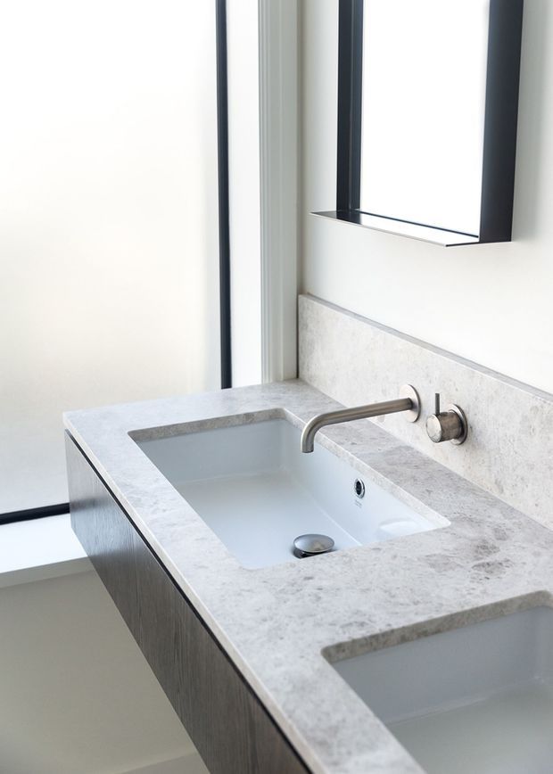 Close-up of a contemporary bathroom vanity with an integrated double basin in a pale stone benchtop, brushed metal wall-mounted tapware, light grey textured splashback, dark timber-look cabinetry, and a large black-framed mirror. Bright, minimal and refined composition.