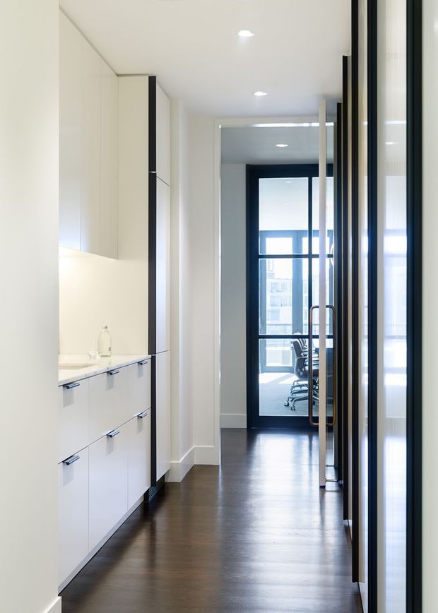 A bright, minimalist interior corridor with full-height white built-in cabinetry and a compact counter along the left wall. Dark timber flooring contrasts with the white joinery and walls, while black-framed glazed doors and partitions line the right side and lead toward a glass-fronted office or workspace at the end. Recessed ceiling lighting and clean, flush detailing create a refined contemporary feel.