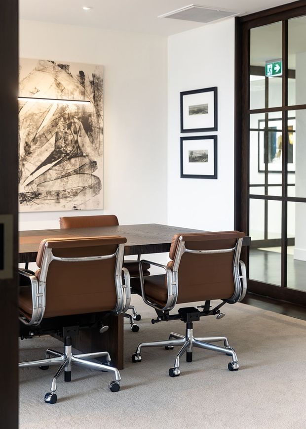 A refined office-style dining or meeting room with a rectangular dark wood table, four brown leather-and-chrome swivel chairs, pale carpet, white walls, framed artwork, and dark timber-framed glazed doors. The room has a clean, professional mid-century-inspired character with a glass-partitioned space beyond.