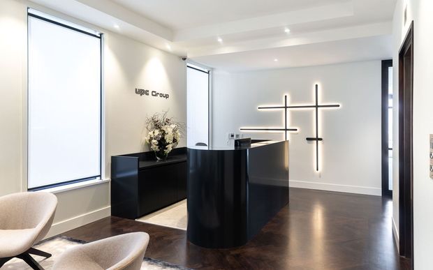 A refined commercial reception area with a glossy black curved reception desk, matching low credenza, upholstered lounge chairs, dark timber flooring, bright frosted windows, and minimalist linear wall lighting. The space has a clean, contemporary corporate aesthetic with high contrast between white walls and dark joinery.