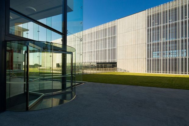 Exterior architectural view of a contemporary institutional or commercial building surrounding a landscaped courtyard. The foreground features a glazed entrance volume with a revolving glass door, dark metal framing, and reflective curtain wall glazing. Across the courtyard is a large rectilinear facade screened by dense vertical fins, with a pale neutral finish and bands of windows. A lawn and broad paved forecourt complete the scene under clear daylight.