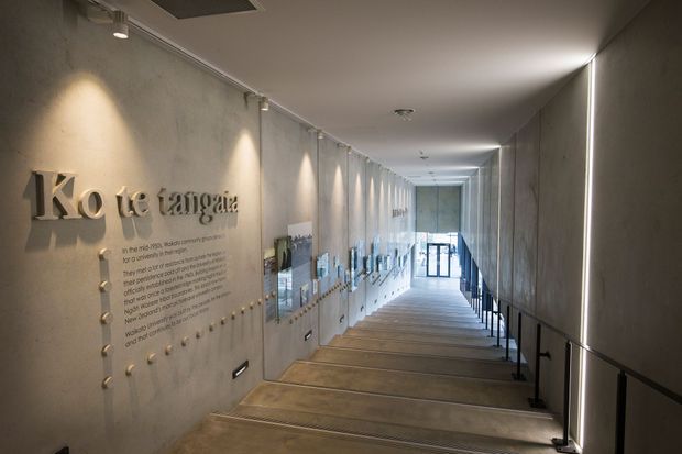 A long, modern interior passage or gallery with a gently descending timber-toned floor, raw concrete walls, black metal handrails, and slim linear lighting. The left wall features large raised lettering and interpretive display panels, while glazing at the far end brings in daylight. The space has a refined institutional or commercial exhibition character.