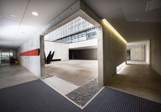 Interior view of a large contemporary institutional or commercial circulation space centred on a raw concrete courtyard-like atrium. Broad concrete walls and floors frame an open void with upper-level glazing and linear metal balustrades. A narrow gravel strip and recessed linear lighting add detail, while dark entrance mats, a red wall feature, and black sculptural forms provide contrast. The space has a minimalist, industrial character with high ceilings and strong geometric lines.