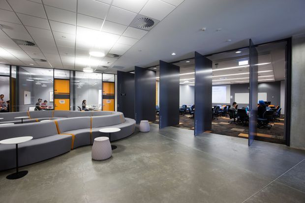 A contemporary commercial office interior with an open reception or breakout lounge in the foreground and glazed meeting or work rooms beyond. The space features modular grey upholstered seating with orange accents, dark blue-black partition walls and door frames, exposed polished concrete flooring, glass walls, a suspended acoustic ceiling grid, recessed and linear lighting, and visible office workstations.