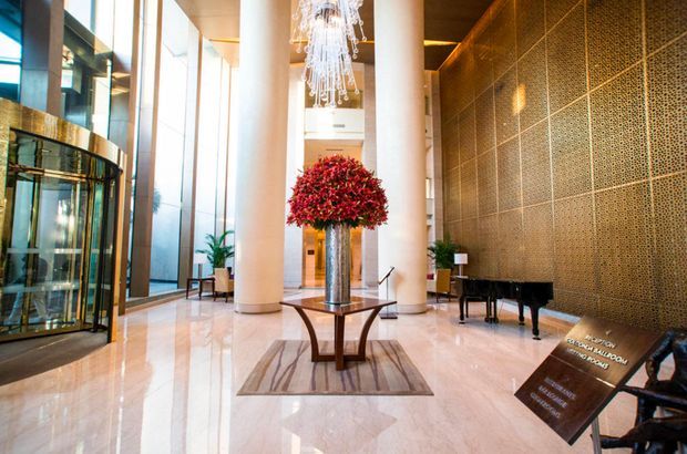 Grand contemporary hotel or commercial lobby with a polished pale stone floor, tall white cylindrical columns, floor-to-ceiling glazing, a dramatic suspended chandelier, a large floral arrangement on a central table, and a textured metallic feature wall with a black grand piano. A revolving glass entrance is visible on the left.