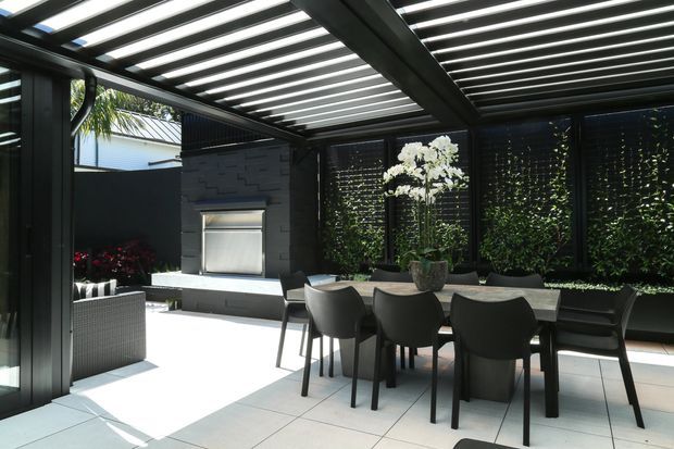 A contemporary covered outdoor dining area beneath a dark louvred pergola. A long dining table with black chairs sits on large pale tiled flooring, beside a dark built-in outdoor kitchen or barbecue wall. Vertical greenery screens and black-framed glazing create privacy, with bright daylight filtering through the overhead slats.