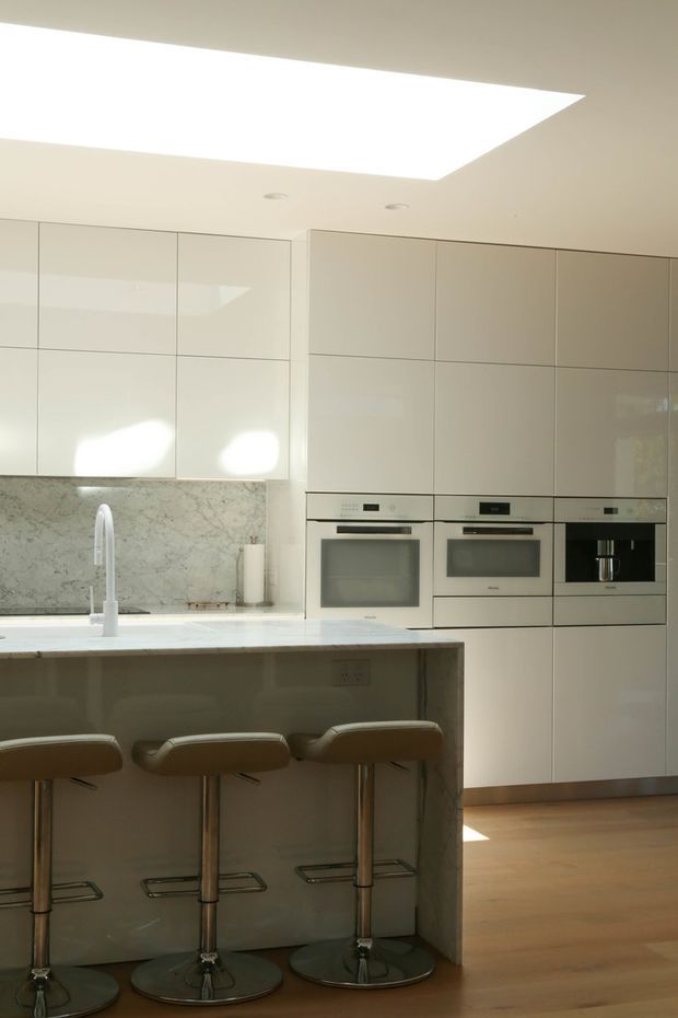Bright contemporary kitchen with a large island and three upholstered bar stools. Full-height glossy white cabinetry integrates double ovens, a microwave, and a coffee machine. A pale veined stone backsplash and island benchtop contrast with warm timber flooring. A large rectangular skylight provides abundant natural light.