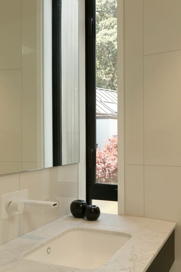 A close-up view of a refined bathroom vanity. The scene features a pale stone benchtop with an integrated white basin, a minimalist white wall-mounted tap, large pale rectangular wall tiles, and a tall black-framed window overlooking greenery and a metal roof. A mirror runs along the left side, reflecting the clean, light-filled space. Small black decorative vessels provide contrast on the vanity.