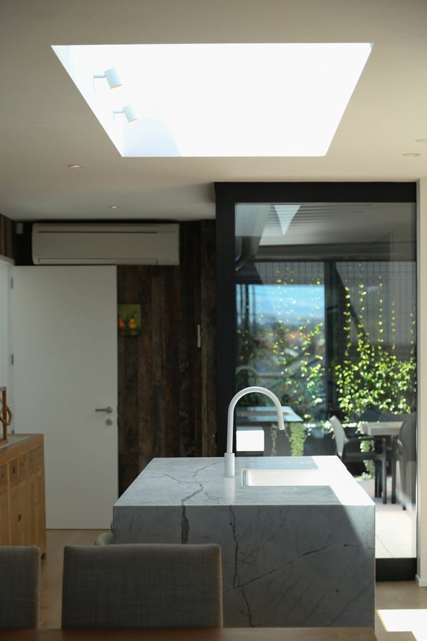 A contemporary kitchen interior centered on a substantial pale veined stone island with an integrated sink and curved gooseneck tap. The island is paired with upholstered grey seating, while dark timber wall panelling, pale cabinetry, a white door, black-framed glazing, and a large rectangular skylight define the space. Sunlight enters through the skylight and rear glazing, creating a bright, refined atmosphere.