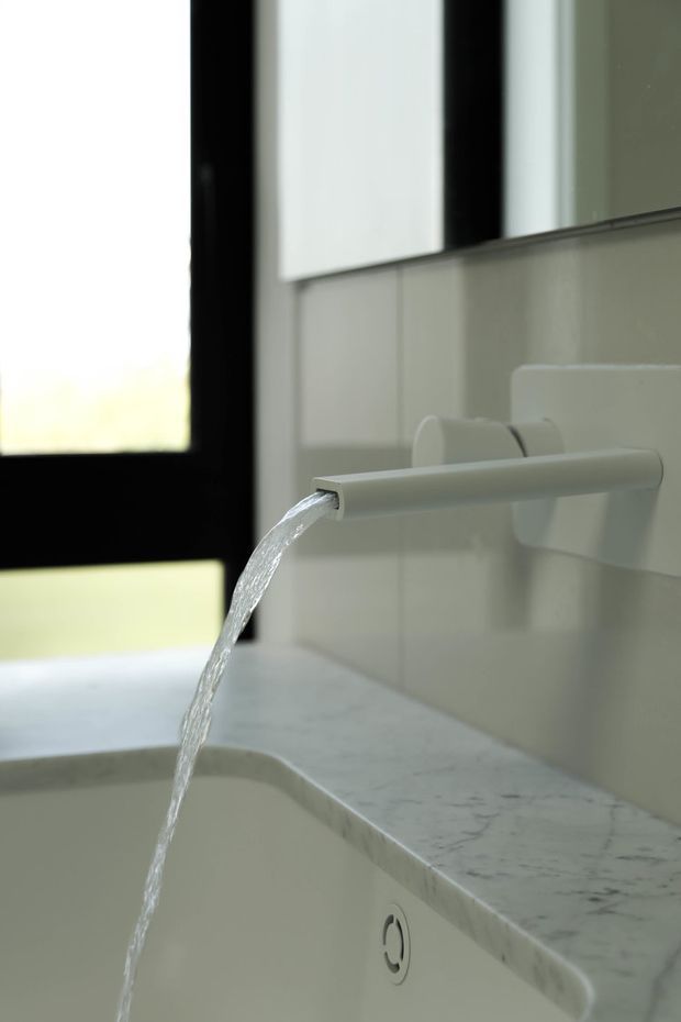 Close-up of a minimalist bathroom vanity or basin with a wall-mounted white cylindrical faucet running water into a curved integrated basin. The surrounding surfaces are pale marble or stone, with light tiled wall finishes and a black-framed window visible at left.