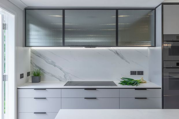 Contemporary kitchen with sleek white handleless cabinetry, a white marble-look splashback and benchtop, dark integrated appliances, and upper cabinets fitted with textured ribbed glass doors in black frames. Under-cabinet lighting highlights the clean, refined workspace.