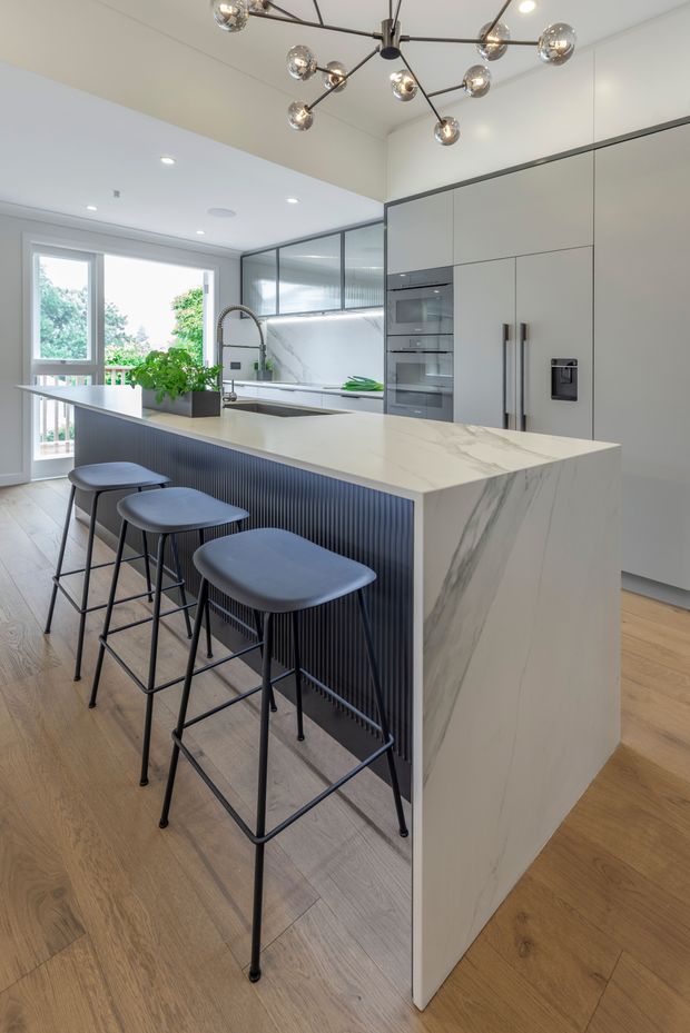 Contemporary kitchen interior with a large white marble-look island, integrated dark fluted panel, three black bar stools, pale timber flooring, streamlined white cabinetry, dark-framed internal glazing, and a sculptural multi-globe pendant light. The space is bright and open, with greenery visible through the rear glazing.