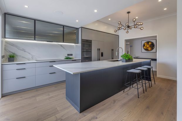 Contemporary kitchen with a large island clad in dark vertical fluting, pale timber flooring, matte light cabinetry, integrated appliances, and a dramatic white marble-look splashback. The island includes seating for four beneath a sculptural branching pendant light, with black-framed glazed cabinetry and a dining or living area visible beyond.