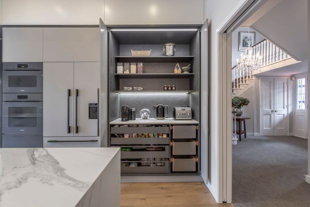 A refined contemporary kitchen viewed toward a built-in appliance wall and open pantry-style shelving. The kitchen features matte light-grey cabinetry, integrated ovens, a large white marble-look island benchtop, dark open shelving with drawers and integrated appliances, and warm timber flooring. Through the doorway, a carpeted entry hall with a white door, staircase, and decorative chandelier is visible.