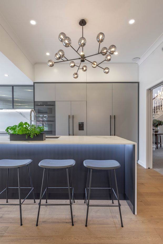 A contemporary kitchen with a large island breakfast bar, three stools, matte grey full-height cabinetry, integrated appliances, and a dark vertically fluted island front. The island has a pale stone benchtop and a black multi-arm globe chandelier above it. Warm timber flooring, recessed ceiling lights, and a glimpse of an adjoining room complete the space.
