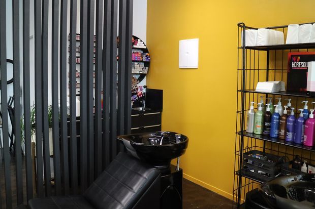 A contemporary hair salon wash area with black shampoo basins and reclining wash chairs in front of a mustard-yellow wall. A black vertical-slat partition partially screens the space, while open black metal shelving displays towels, hair products, and salon supplies.