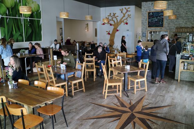 A busy casual café or restaurant interior with numerous wooden tables and chairs, a service counter, chalkboard menu, and several customers. The space features wood-look flooring with a large compass-star inlay, a stone-textured feature wall, white walls, timber ceiling panels, pendant lighting, and botanical mural artwork.