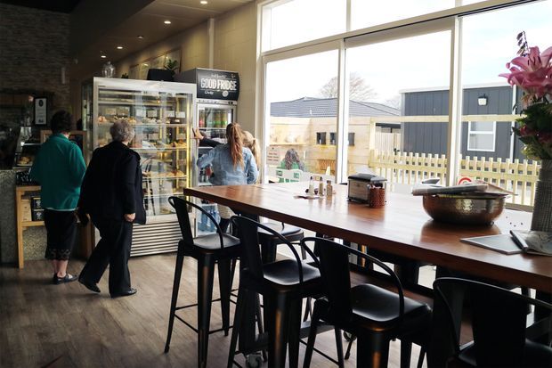 A casual café interior with a long communal timber table and black bar stools beside large floor-to-ceiling windows. A refrigerated display cabinet, counter area, customers, and food-service fixtures occupy the darker left side, while an outdoor courtyard and neighbouring buildings are visible through the glazing. The space has warm timber flooring, dark ceiling surfaces, and a relaxed contemporary hospitality feel.
