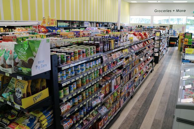 Bright interior view of a grocery supermarket aisle, with long stocked shelving displaying packaged food and canned goods, refrigerated displays along the right side, and a light striped feature wall near the checkout or store entrance. The space has a clean, contemporary retail fit-out with pale timber-look flooring and bright overhead lighting.