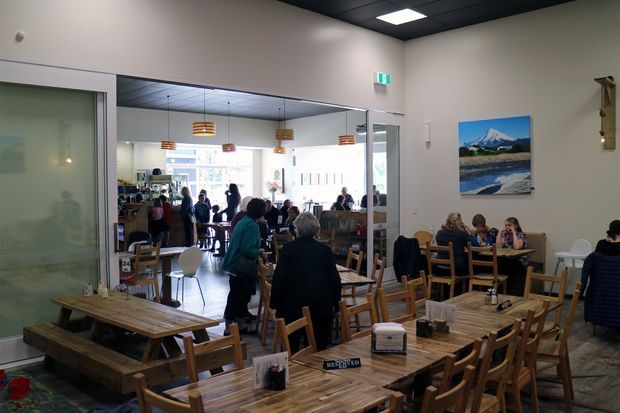 A bright casual café dining area with numerous timber tables and chairs, communal bench seating, pendant lights, and customers seated throughout. The space has a modern suspended ceiling, pale walls, large glazed partitions, and a mountain landscape artwork on the wall.